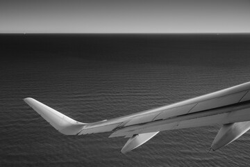 A high-quality black-and-white photo captures a striking perspective from an airplane wing, with the sea below and the sky above the horizon. The contrast creates a serene and dramatic atmosphere.