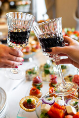 Two hands clinking crystal wine glasses filled with red wine, set against a blurred background of appetizers on a table. The image captures a celebratory toast at a social gathering or event.