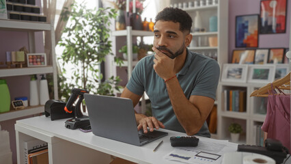 Young man looking thoughtful in a home decor store, seated at a desk with a laptop and various product packaging tools and surrounded by stylish decorations.