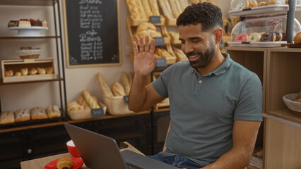 Fototapeta premium Young man making a video call inside a bakery, smiling and waving while seated at a table with a laptop and pastry