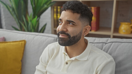 Fototapeta premium Handsome bearded man relaxing on couch with bookshelf and plant decor in cozy living room.