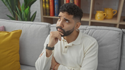 A thoughtful young hispanic man with a beard sits contemplatively in a cozy living room, implying introspection