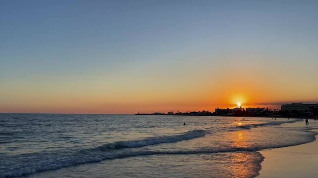 Sunset over the sea in Ayia Napa, Cyprus, slow motion with gentle waves and people swimming. In the distance, a city silhouette adds peaceful autumn scene during the travel velvet season.