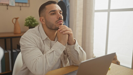 Naklejka premium A pensive young hispanic man at home with a laptop in a modern apartment, reflecting a contemporary lifestyle.