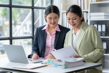 Fototapeta premium Collaborative Success: Two professional women in a modern office, engaged in discussion over documents, laptops, and data, symbolize the power of partnership and teamwork in achieving shared goals. 