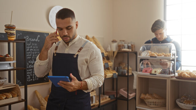 Men working in a bakery, one taking a phone call and managing an electronic tablet while the other arranges pastries in a modern, well-lit shop interior