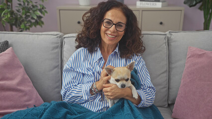 A smiling middle-aged woman cuddles a chihuahua on a cozy sofa indoors, showcasing companionship and comfort at home.