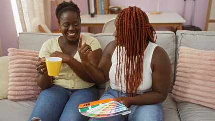 Two african american women enjoying quality time together in a cozy living room, one holding a coffee cup and the other holding color samples with smiles and laughter