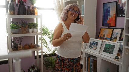 Mature woman with grey hair and glasses smiling while talking on mobile phone and holding papers in a colorful home office with pink wall background