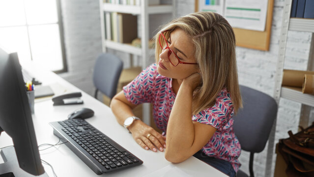 Woman working at computer in office, wearing glasses, feeling neck pain, seated at desk with keyboard and mouse, indoor setting with shelf and documents in the background - Powered by Adobe