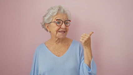 Elderly woman with grey hair and glasses gesturing thumbs up against a pink background