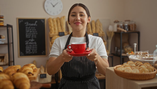 Smiling young hispanic woman holding red cup in a cozy bakery shop with bread and pastries in the background