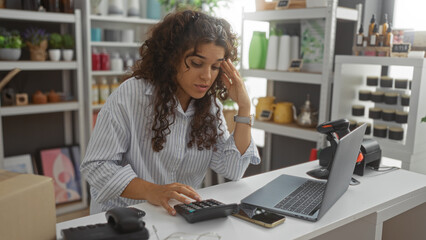 Woman calculating finances in a decorated store with shelves of products behind her, using a laptop and calculator at the counter