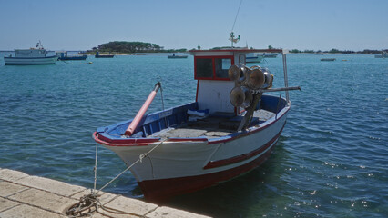 Obraz premium Fishing boat docked in the clear blue waters of porto cesareo, italy, with other boats and an island in the background under a bright sunny sky.