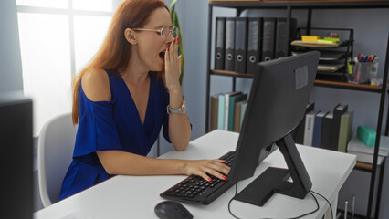 Young woman yawning at desk in office room with shelves of books and folders while working on...