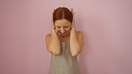 Young woman with red hair covering her ears due to noise over isolated pink background