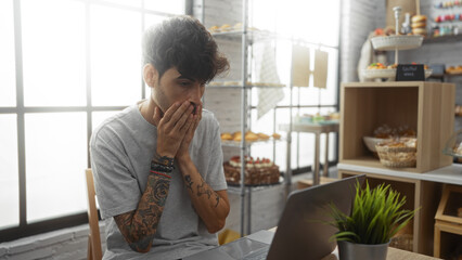 Young man with a beard and tattoos looking surprised while sitting in a bakery cafe with a laptop on the table