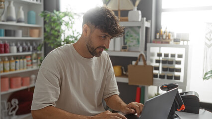 Young man working in a home decor store, surrounded by various decorations and plants, focused on his task at the laptop