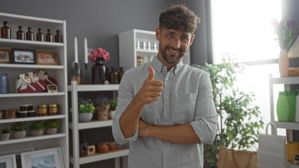 Handsome young man giving thumbs up in a modern home decor shop with shelves filled with various decorative items and plants in the background.