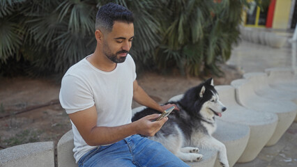 Hispanic man with smartphone sitting beside husky dog in urban park.