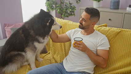 A young hispanic man relaxes at home, bonding with his husky dog on a yellow couch, holding a coffee mug. © Krakenimages.com