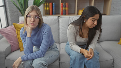 Two women sitting on a couch in a living room look pensive and sad, reflecting a somber mood at...