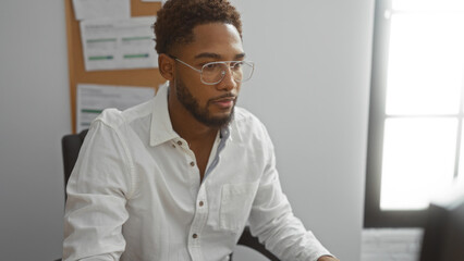 Fototapeta premium Handsome man working in an office setting, focused on a computer screen, wearing glasses and a white shirt, with a noticeboard and window in the background