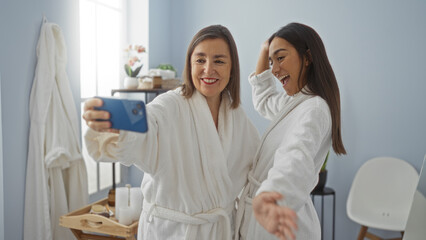 Mother and daughter in white bathrobes taking a selfie together, smiling and enjoying their time in a spa wellness center with light blue walls and minimalistic decor.