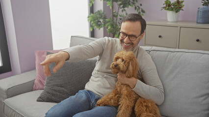 A middle-aged hispanic man joyfully points while holding his poodle in a cozy living room setting,...