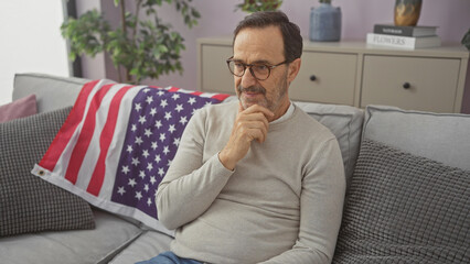Thoughtful mature hispanic man sitting in a living room with an american flag on the couch, contemplating indoors.