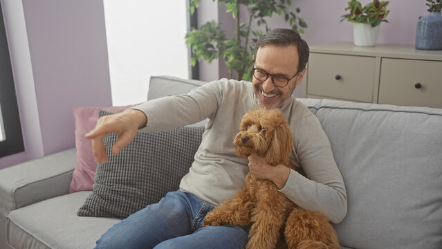 A middle-aged hispanic man joyfully points while holding his poodle in a cozy living room setting, capturing a warm moment at home.