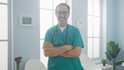 Fototapeta premium A handsome hispanic male doctor in green scrubs stands confidently with crossed arms in a bright clinic lobby with white chairs, framed artwork, and a wall clock.