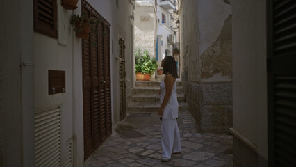 A young hispanic woman in white clothing explores the charming streets of polignano a mare in puglia, italy, surrounded by historic buildings and potted plants.