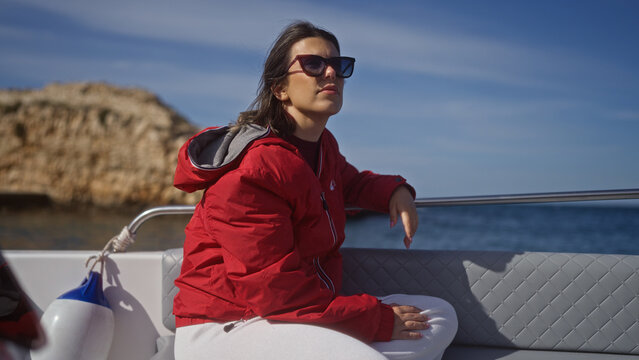 Young hispanic woman wearing sunglasses and a red jacket, seated on a boat in polignano a mare, puglia, italy, enjoying the serene sea and scenic cove under a bright, clear sky.