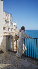 A beautiful young hispanic woman enjoys a scenic view at a viewpoint in polignano a mare, puglia, italy, overlooking the blue sea and historic architecture.