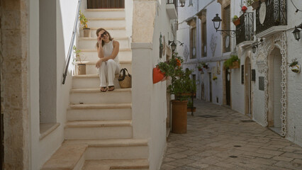 A young beautiful hispanic woman sits on white steps in the charming old streets of locorotondo, puglia, italy, basking in the european allure of this picturesque town.