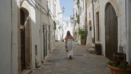 Young hispanic woman walking in the charming old town of ostuni, puglia, italy, europe, wearing a white dress and holding a straw bag in a narrow ancient street with whitewashed buildings.