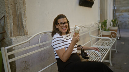 Young hispanic woman enjoying ice cream while sitting on a bench in lecce, a charming town in puglia, italy, showcasing her beauty and the serene ambiance of the italian street.