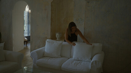 Young hispanic woman arranging cushions on a white sofa in a beautifully lit italian masseria...