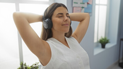 Woman relaxing with headphones in an office environment, showcasing a serene moment, young, adult, female, attractive, brunette, indoors with a hint of indoor plants, organized workspace.