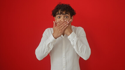 A surprised hispanic man in a white shirt covers his mouth against a vibrant red background, conveying a strong emotional reaction.