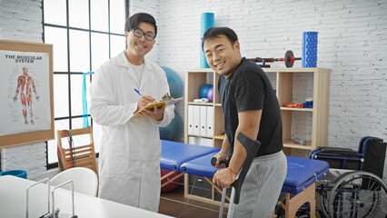 Obraz premium A smiling asian doctor with a clipboard and a male patient with crutches in a well-equipped physiotherapy clinic.