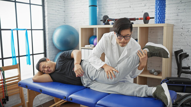 Physiotherapist male assisting another man doing leg exercise in a well-equipped physiotherapy clinic.