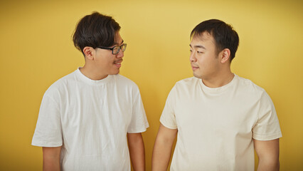 Fototapeta premium Two asian men in white shirts engage in conversation against a plain yellow background, depicting friendship or casual interaction.