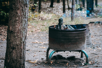 A smoldering fire in a rustic metal hearth with charred windows giving off faint smoke. The outdoor...