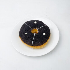 A round donut covered in dark crumbs, topped with white icing lines and dots, served on a clean white plate. The donut’s rich topping contrasts with the light background.