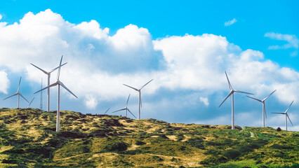 Wind turbines are situated on a grassy hill, with a large white cloud overhead.