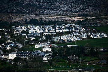 A panoramic view of a town nestled amidst rolling hills. The town features a mix of residential buildings with different sizes and colors. There is a visible winding road leading through the town.