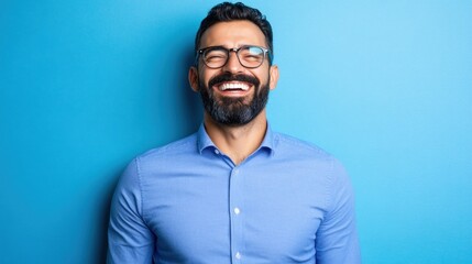 Fototapeta premium A bearded man with glasses is smiling widely, exuding happiness and confidence, standing against a vivid blue backdrop in a casual setting