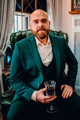 person in green blazer sits on a leather sofa, holding a wine glass. background features a blurred fireplace, creating a cozy and elegant atmosphere. Perfect for lifestyle and interior design themes.